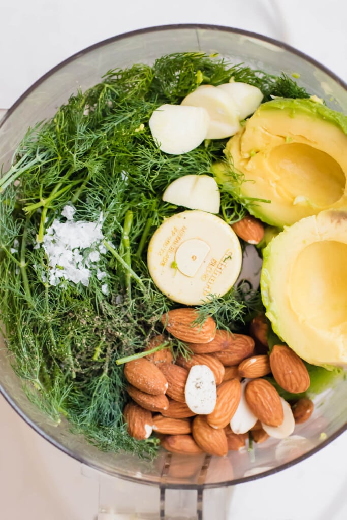 the bowl of a food processor filled with dill, avocado, garlic, and almonds
