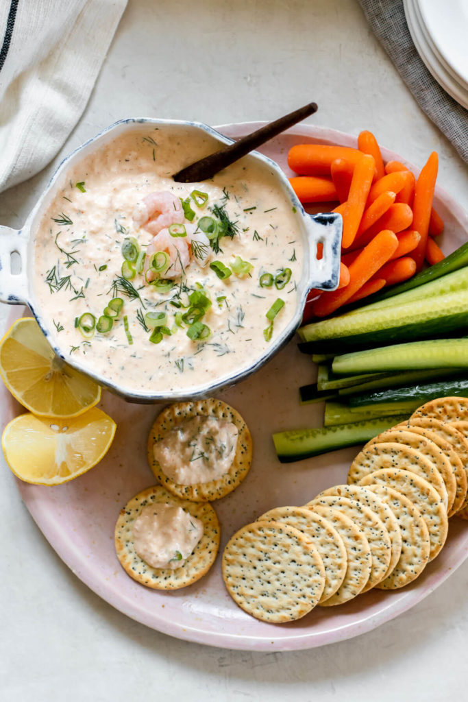 large platter of veggies and crackers with a blue colored bowl filled with shrimp dip