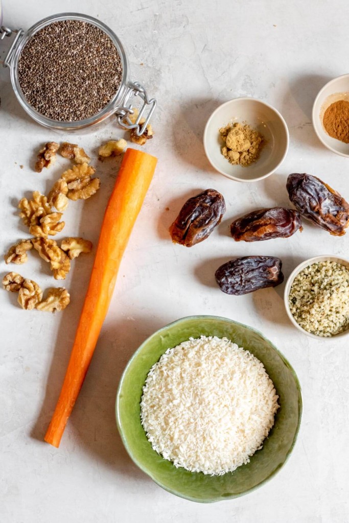 Ingredients needed for carrot cake bites on white background, carrots, dates, walnuts, coconut, hemp seeds, spices, and chia seeds