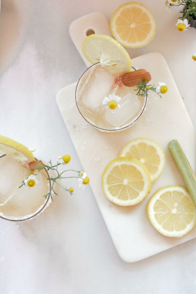 2 glasses filled with rhubarb chamomile margarita and garnished with lemon and rhubarb on a white background with lemon slices
