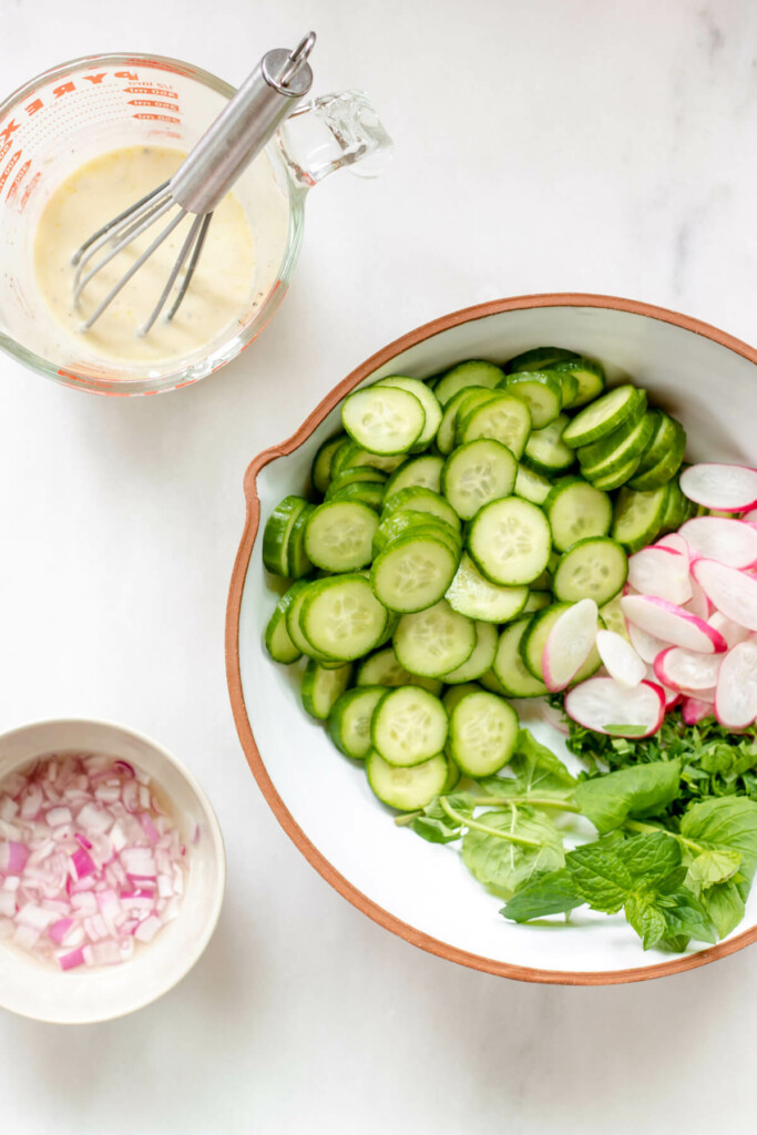 Bowl of cucumbers, radish, and herbs, and a small white bowl of onions and a pyrex bowl of creamy yogurt dressing