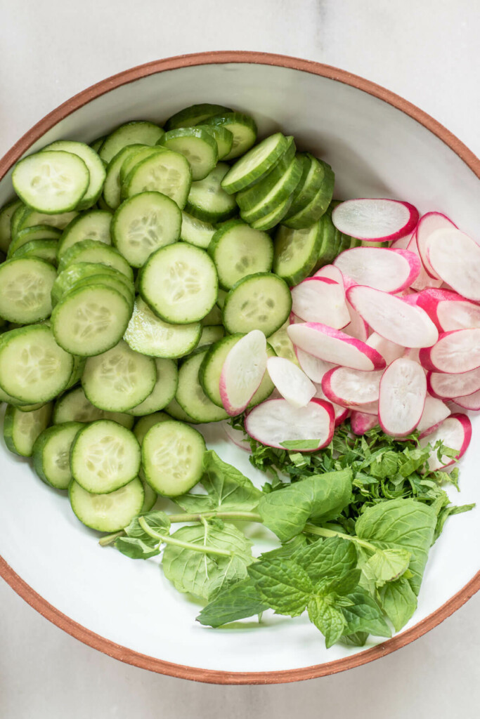 close up shot of cucumbers, radish, chives, and mint in a white bowl on a white background