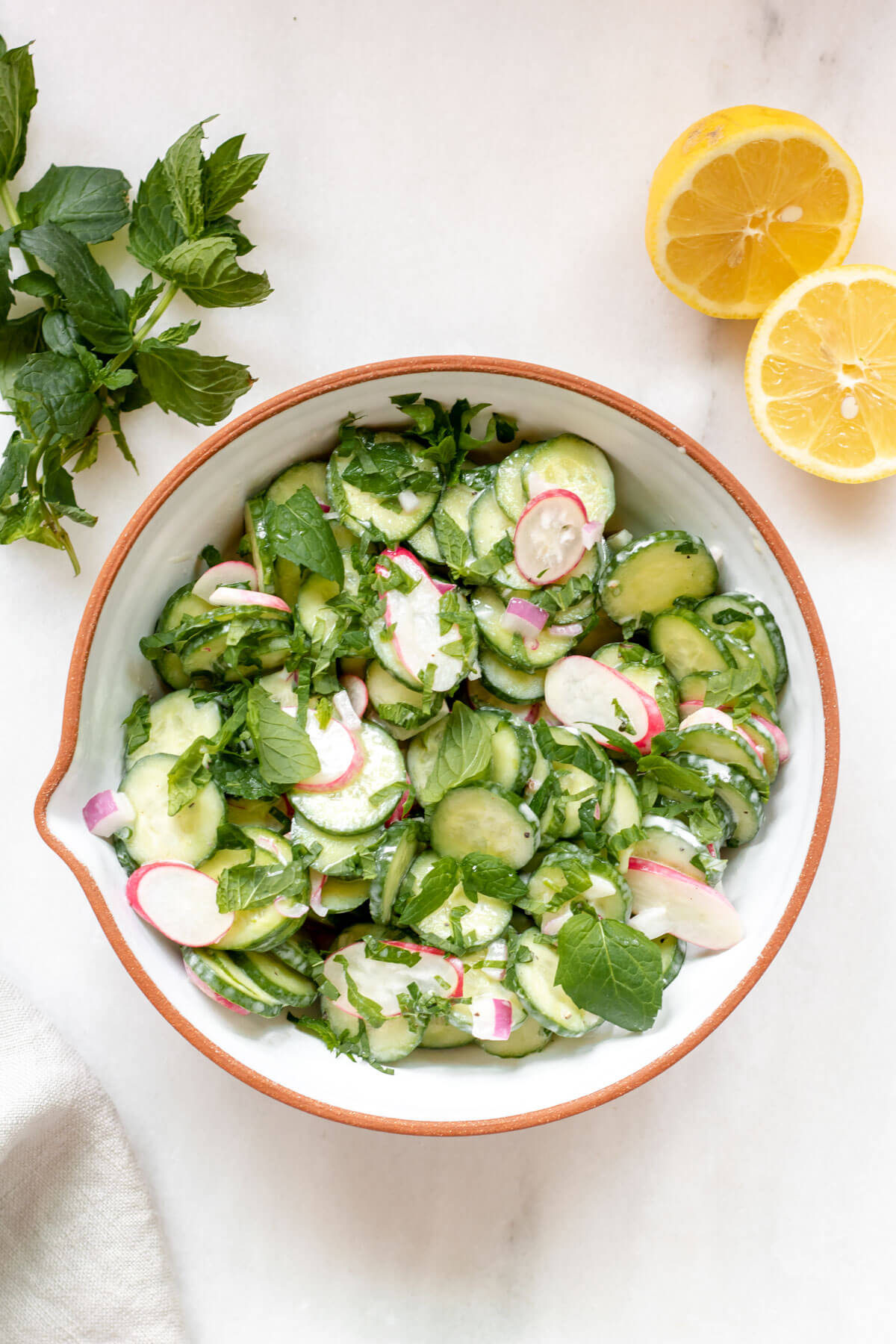 white bowl of cucumber radish salad on white background with lemon and mint in the background