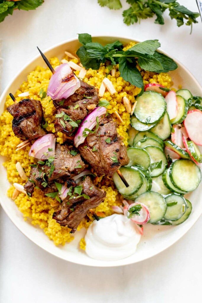 grilled lamb kebabs served over turmeric quinoa with cucumber salad. on a white background with lemon and mint on the table