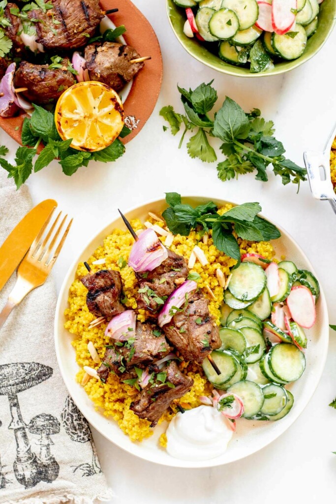 grilled lamb kebabs served over turmeric quinoa with cucumber salad. on a white background with lemon and mint on the table