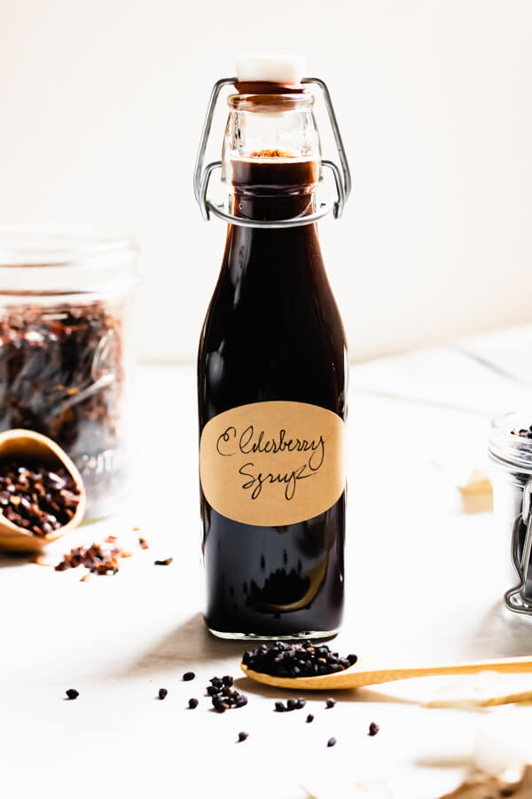 Glass jar of elderberry syrup on white background with scattered elderberries in background