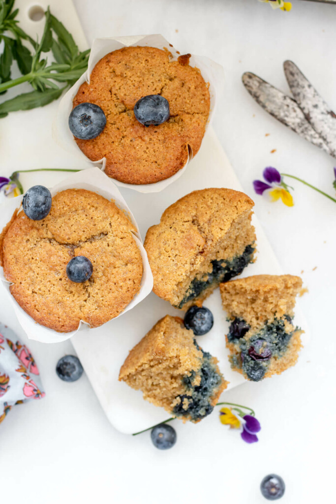 3 gluten free blueberry muffins on a white background with 2 knives and one muffin cut open