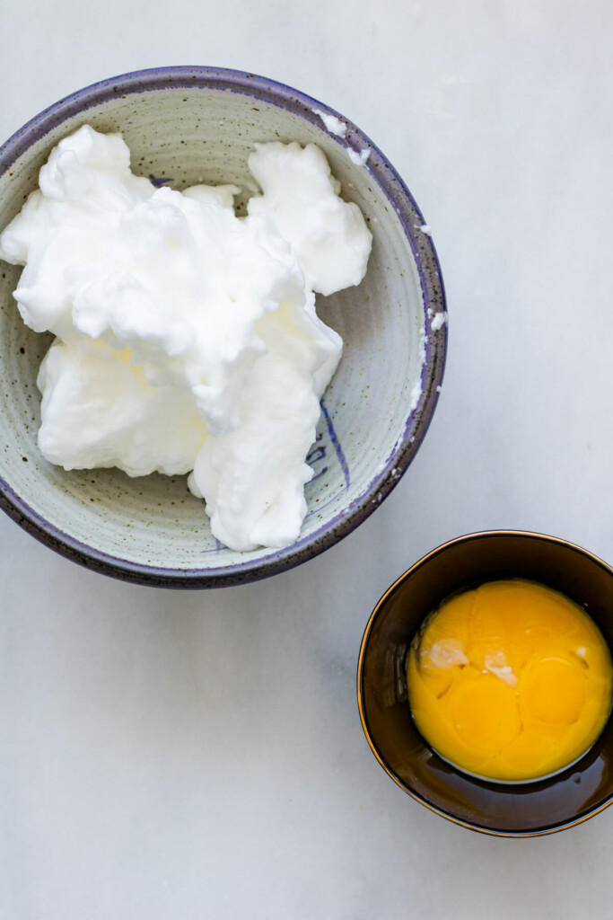 A photo of whipped egg whites in a bowl and a separate bowl of egg yolks on a white background