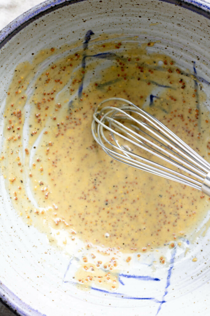 a bowl with mustard, vinegar, and olive oil, and a whisk whipping together the dressing for a no mayo ramp potato salad