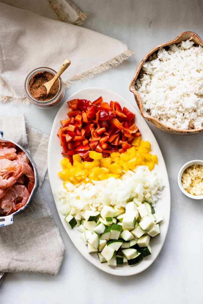 a large platter of vegetables, a bowl of shrimp, a bowl of rice, and cajun rice. all ingredients for cajun shrimp and rice on a white background