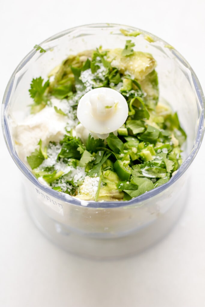 photo of a bowl from a food processor filled with ingredients to make avocado cream, avocado, cilantro, jalapeno, salt, and sour cream