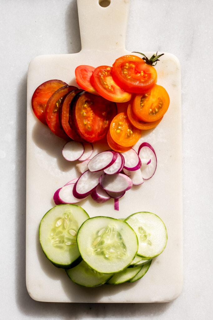 A white marble board with sliced tomato, cucumber, and radish