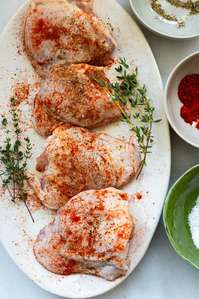 raw chicken thighs on a plate seasoned with paprika, salt, and pepper