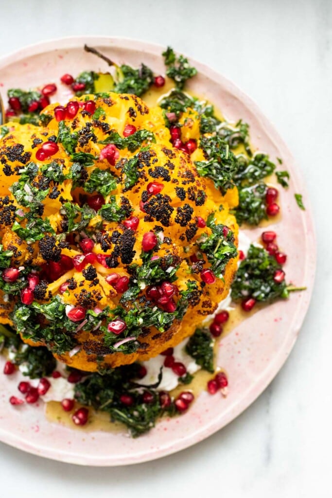 whole roasted cauliflower on plate with green herb sauce and pomegranate seeds