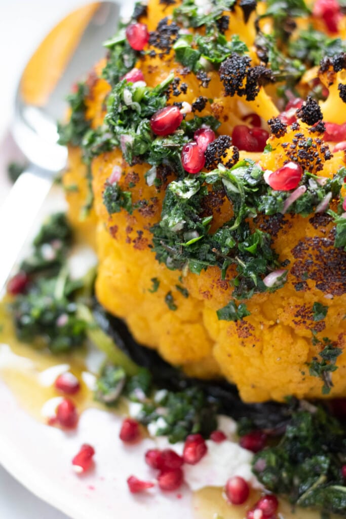 whole roasted cauliflower on plate with green herb sauce and pomegranate seeds