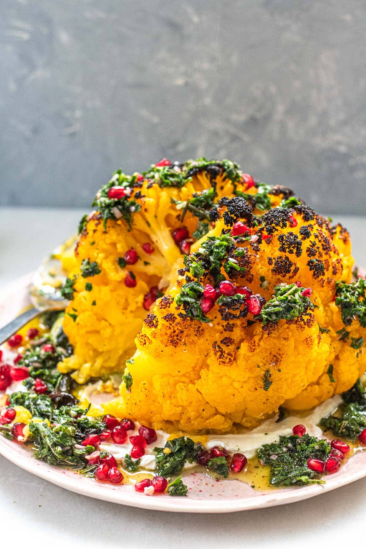 whole roasted cauliflower on plate with green herb sauce and pomegranate seeds