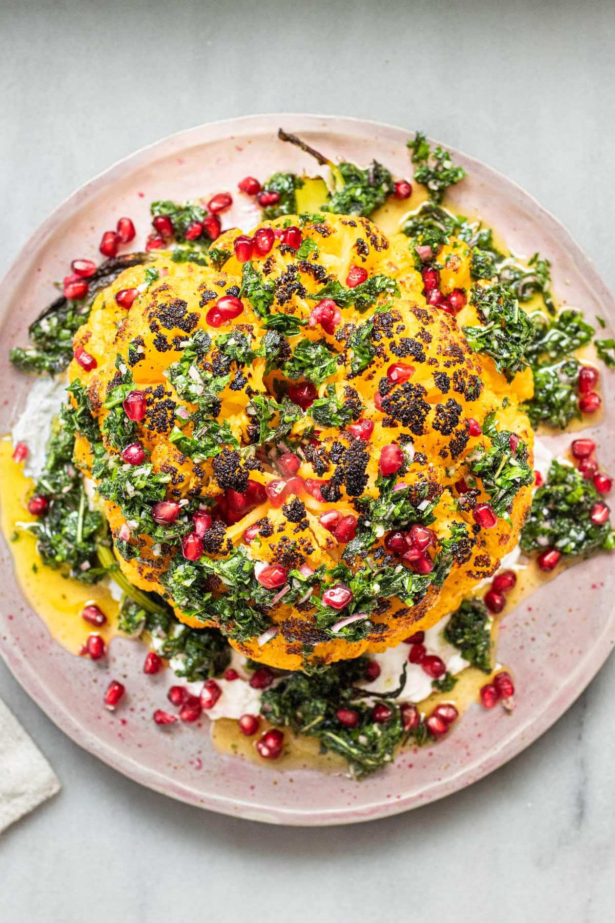 whole roasted cauliflower on plate with green herb sauce and pomegranate seeds