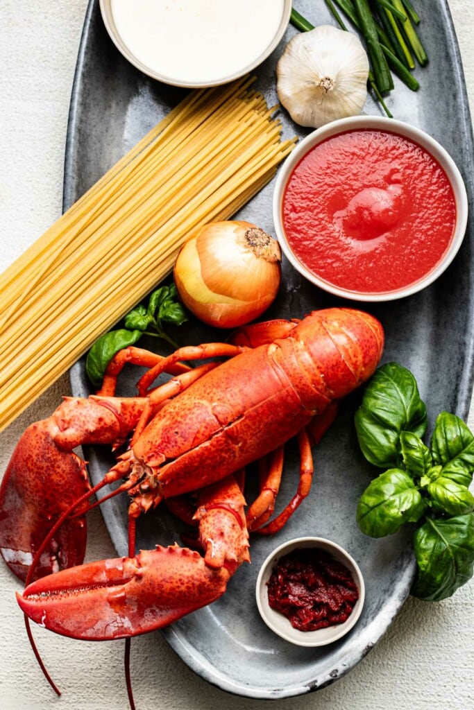 Grey platter of ingredients for lobster pasta including a whole steamed lobster, tomato paste, tomto pasata, onion, garlic, chives, pasta, heavy cream and basil