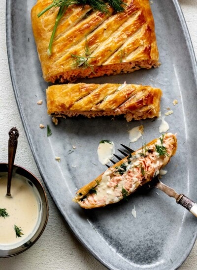 a slice of salmon wellington on a fork with creamy dijon sauce drizzled on top on a grey serving platter