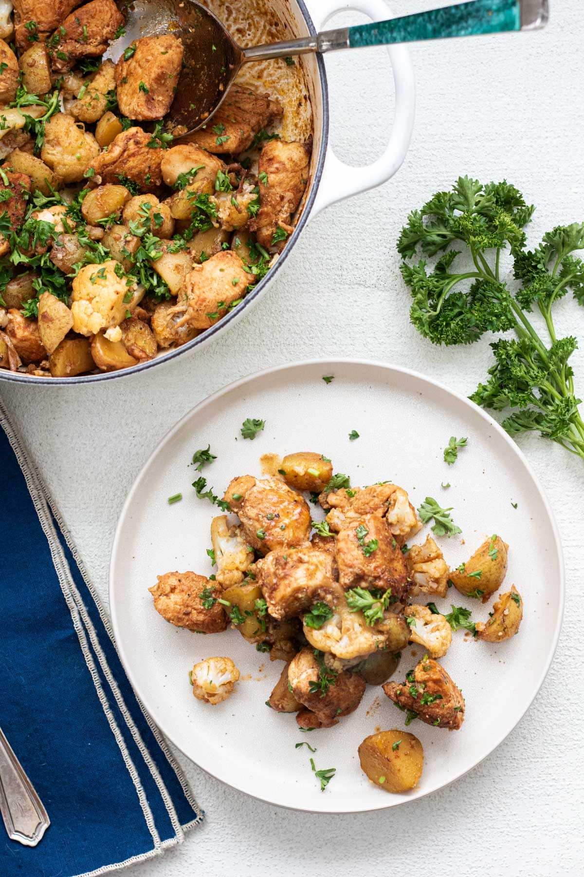 large white pot of chicken and potatoes with a white plate of chicken and potatoes with parsley