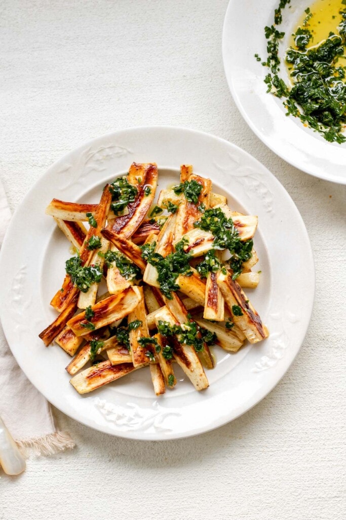 a large white plate piled high with toasty brown roasted parsnips topped with herby green chive gremolata, with a small bowl of herby gremolata in the background