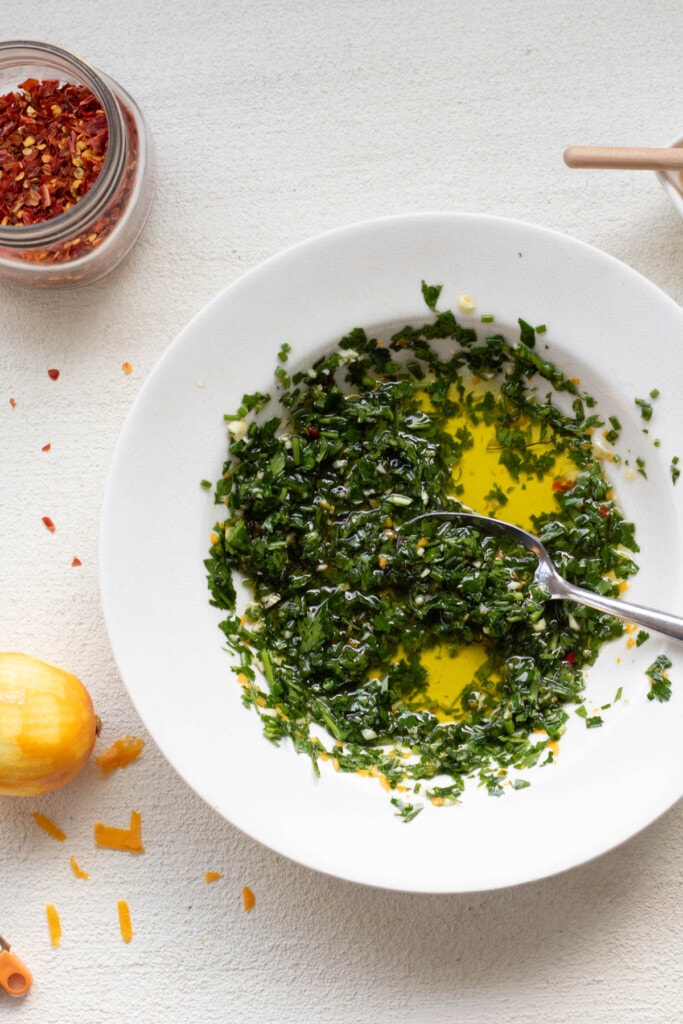 A white bowl with chive gremolata inside and lemon zest on the side with crushed red pepper