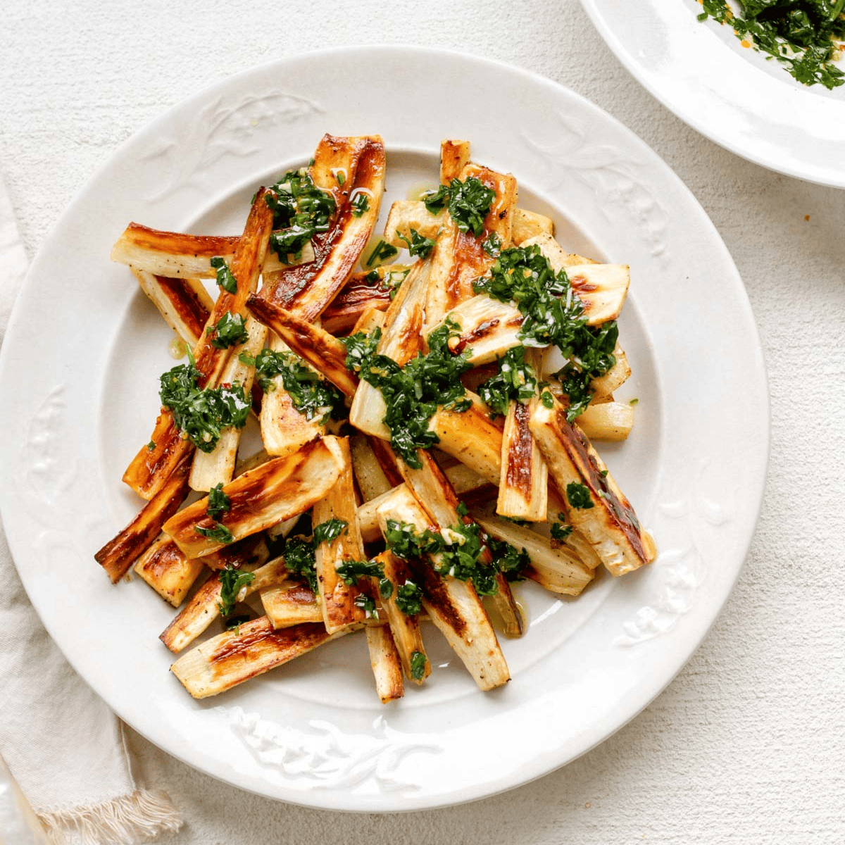 a large white plate piled high with toasty brown roasted parsnips topped with herby green chive gremolata, with a small bowl of herby gremolata in the background