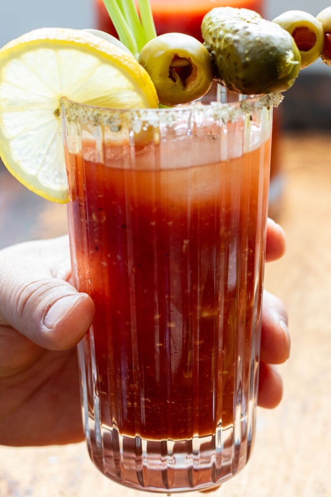 hand holding one glass of bloody mary with pickle, olive, celery, and lemon
