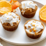 three bakery-style, gluten-free orange flavored muffins on a plate with orange slices and orange peel