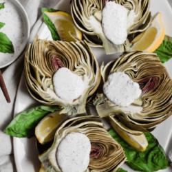 artichokes resting on top of basil leaves on an oval dish beside a creamy sauce