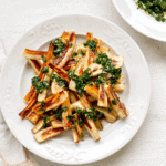 a large white plate piled high with toasty brown roasted parsnips topped with herby green chive gremolata, with a small bowl of herby gremolata in the background