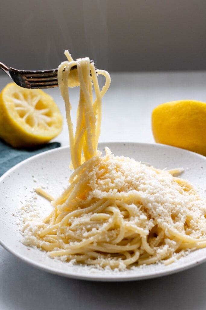 a fork pulling long cheesy lemony noodles in a white bowl