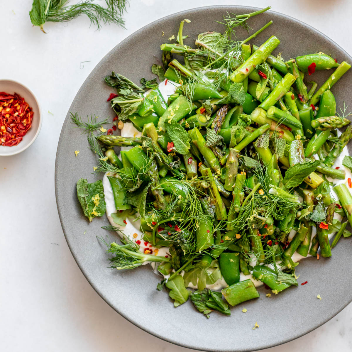 A large grey plate with sugar snap peas and asparagus and a small bowl of crushed red pepper