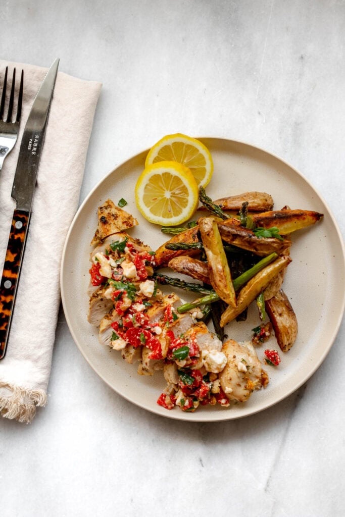 A plate of greek chicken and potatoes with a lemon wedge and fork.