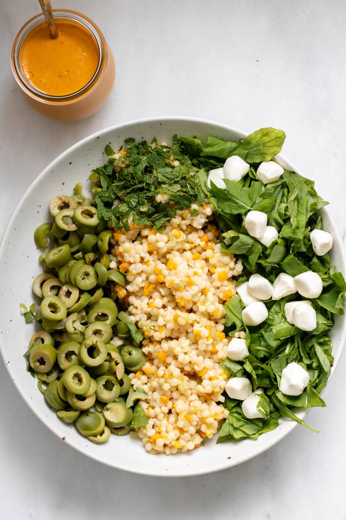 ingredient clusters for olive couscous salad in a white bowl with a jar of tomato vineigrette