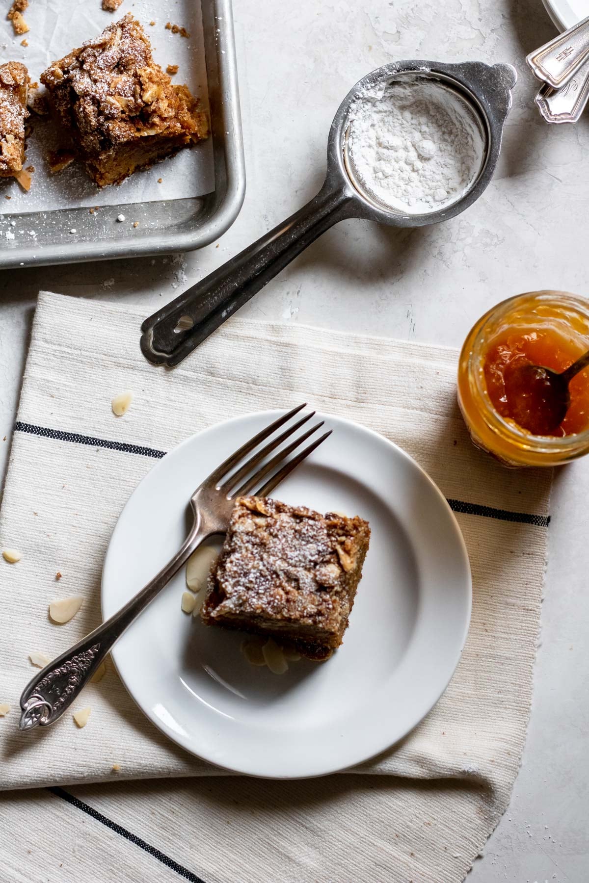 a single slice of Apricot Almond Coffee Cake on a small white plate with a fork and apricot jam in a jar