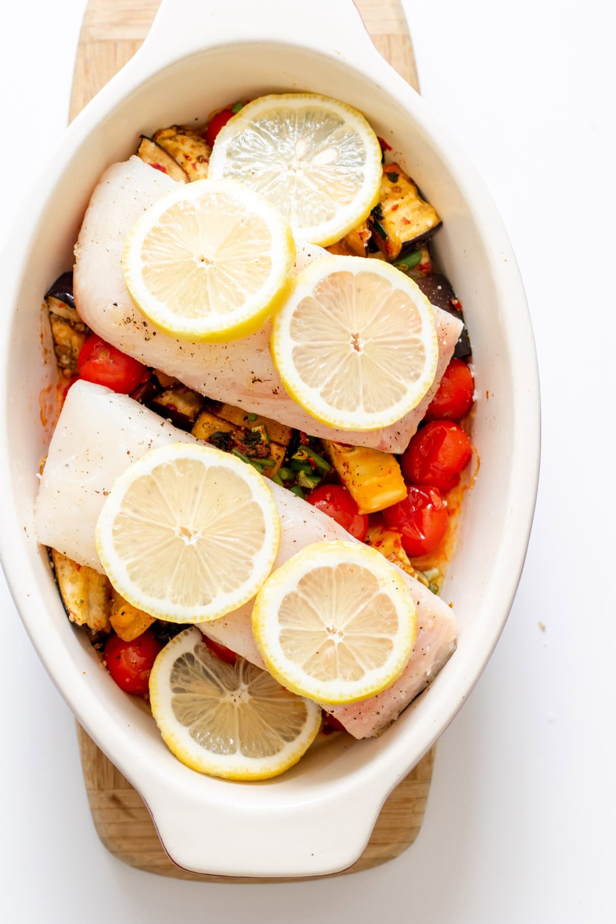 lemon slices on top of raw halibut slices, on top of veggies in a white baking pan